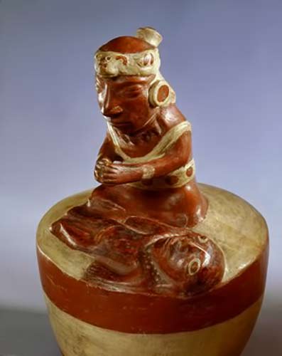 Mocha---shaman-in-prayer Mochica style, this modelled ceramic shows a priest or shaman engaged in a curing ritual or praying over a deceased person. The shaman wears a feline headdress and large disc earrings.