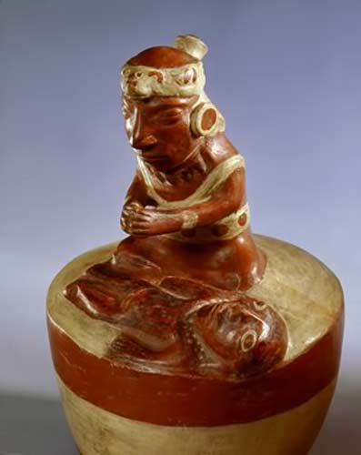 Ceramic-Mochica-Shaman-praying Mocha---shaman-in-prayer Mochica style, this modelled ceramic shows a priest or shaman engaged in a curing ritual or praying over a deceased person. The shaman wears a feline headdress and large disc earrings.