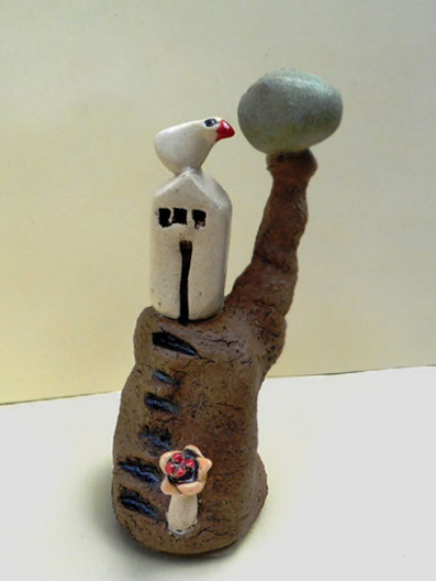 ednapio-etsy A miniature ceramic house on a brown clay hill with a ceramic bird, a tree and a tiny flower.