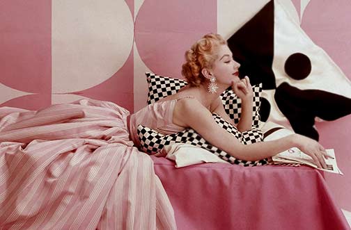Model Lisa Fonssagrives lounging on bed, wearing Claire McCardell's pink and white striped dress. Photo by Richard Rutledge